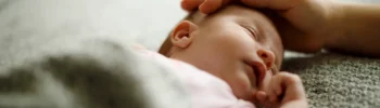 Baby liegt schlafend auf einer hellen Decke. Eine Hand streichelt über seinen Kopf.