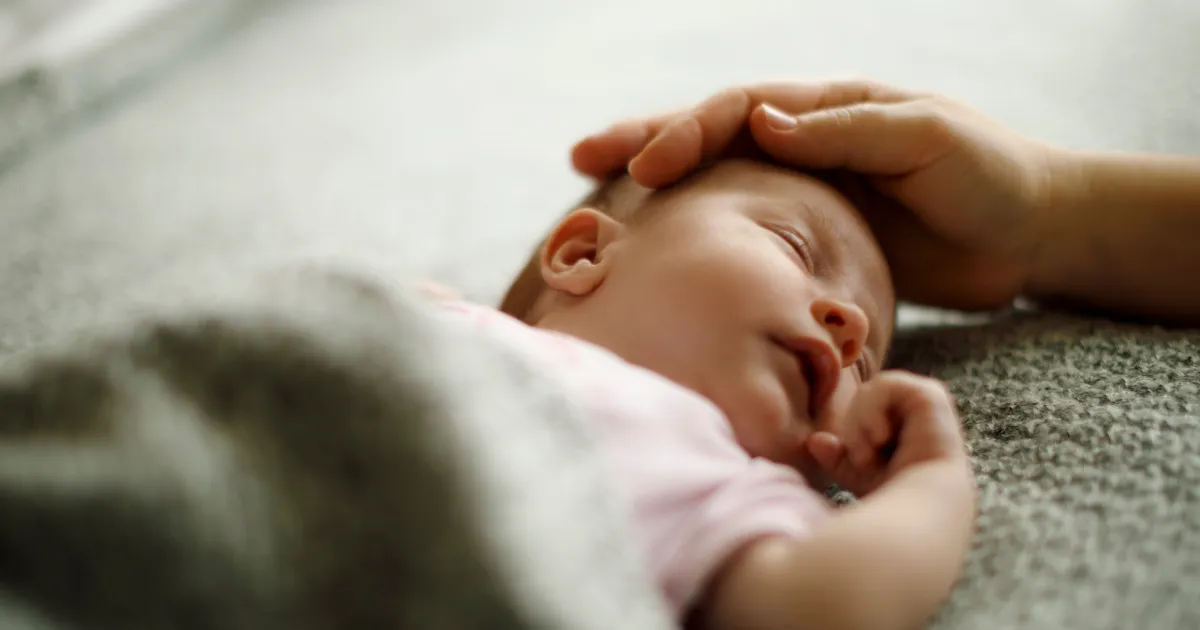 Baby liegt schlafend auf einer hellen Decke. Eine Hand streichelt über seinen Kopf.